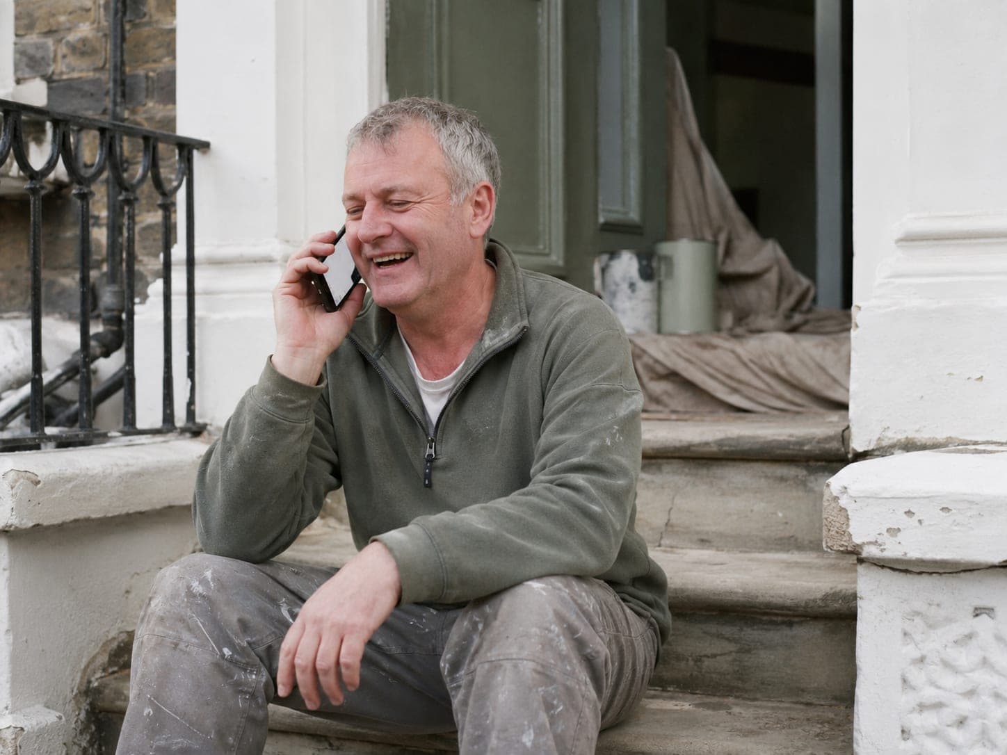 A man in work clothes sitting on the front steps of a house, laughing on the phone.