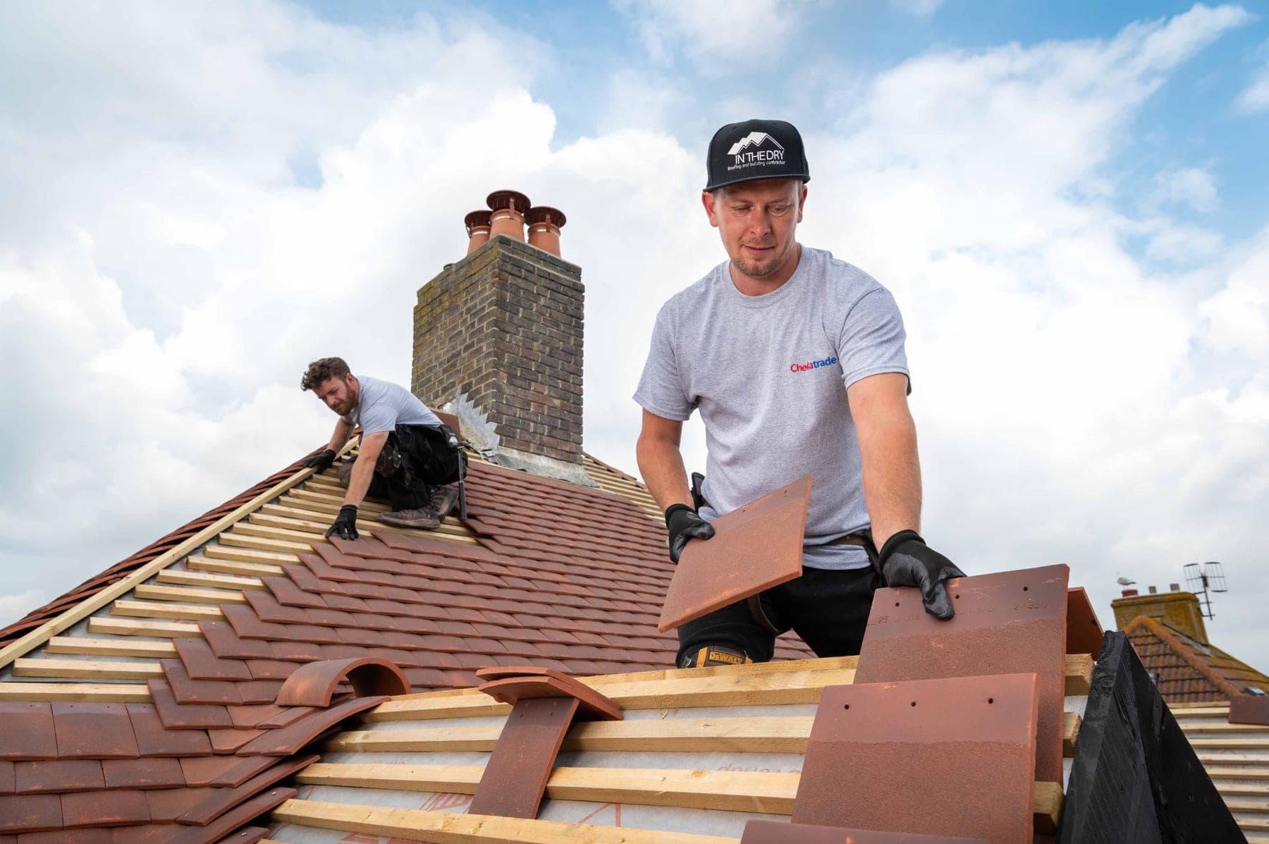Two roofers laying clay tiles on a pitched roof under a bright sky.